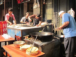 One man stands near a huge pot while three other men prepare food for cooking.