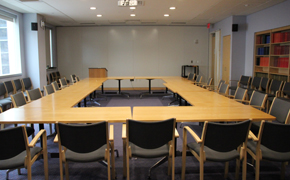 Room with 10 tables arranged in a rectangle, chairs, and a podium. A bookshelf with books along the right wall. A large window on the left wall.