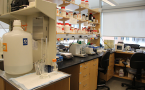 Lab space featuring workbench, chair, and shelving. White bottles with orange caps on the shelves.