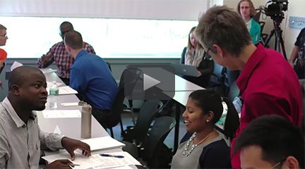 Small groups of students talking at tables in a classroom. In the foreground, an instructor stands near a pair of students talking to each other.