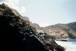 In the left foreground, a rocky hill with sparse patches of shrubs slopes to meet the water. In the background, other hills and outcrops are visible.