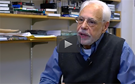 A photo of course instructor Professor David Thorburn seated in his office as he discusses the course. There are shelves behind him cluttered with assorted VHS tapes and books.