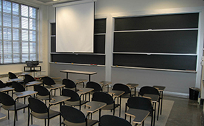 Classroom with about 20 tablet desks arranged in 4 rows. An instructor’s table is at the front of the room. A white projection screen is pulled down in front of a blackboard behind the instructor’s table.