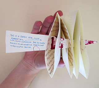 Photo of a hand holding a folded piece of paper with another strip of paper piercing through it to form a locked letter. The paper lock reads: This is a paper lock, now a papered seal with signet impression. The sealing wax acts as an adhesive.