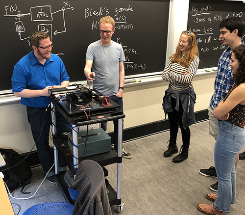 A group of people gathered around an electromechanical device at the front of a classroom.