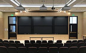 The front of a lecture hall, showing several rows of seats and a set of sliding chalkboards.