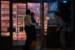 The man with wearing the tie holds a broom while talking to the woman in front of the convenience store. The other man is leaning forward and looking at them.