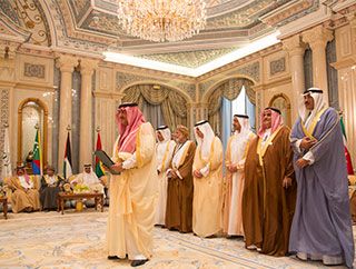 In the middle of a wide-open, elaborate room, six men covered in fabric from head to toe stand behind a similarly-garbed gentleman who holds a binder.