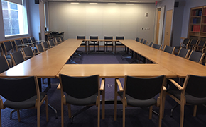 Nine tables arranged in a large rectangle. Chairs arranged along the outside of the rectangle. On one wall, a window. One the opposite wall, book shelves.