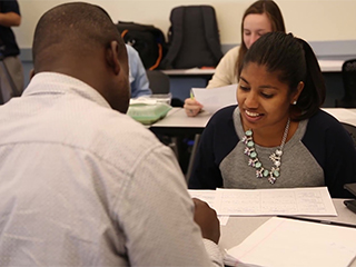 Photo of part of a classroom where 2 students are talking and more students behind them.